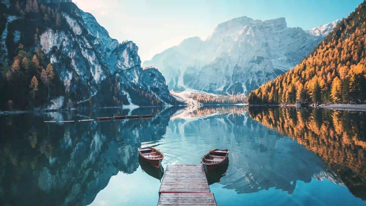 Crystal-clear alpine lake with wooden boats, mountain reflections, autumn forests, and snow-capped peaks at sunrise