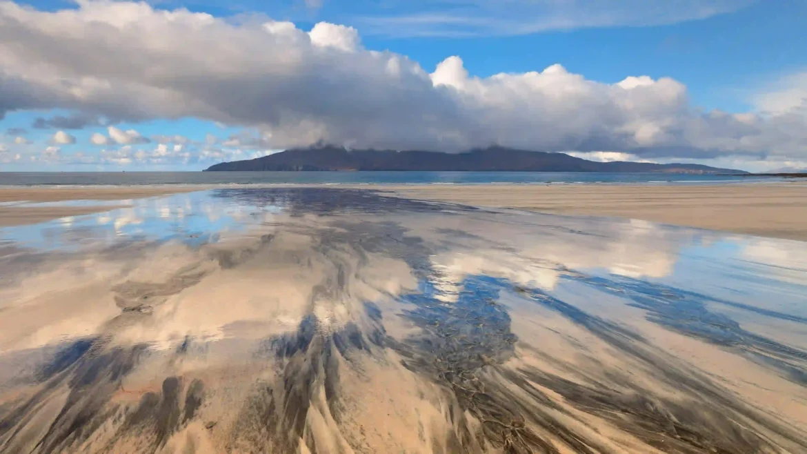 Wide view of a wet sandy beach reflecting blue sky and white clouds with a mountain in the distance 4K nature wallpaper