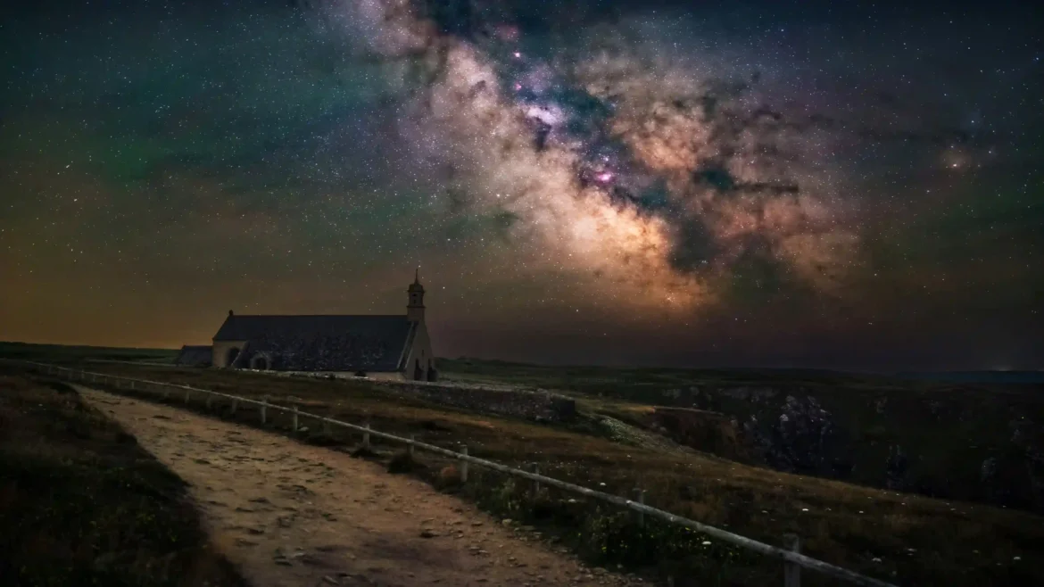 Milky Way galaxy arching across a starry night sky above a small stone chapel on a coastal cliff in a 4K wallpaper