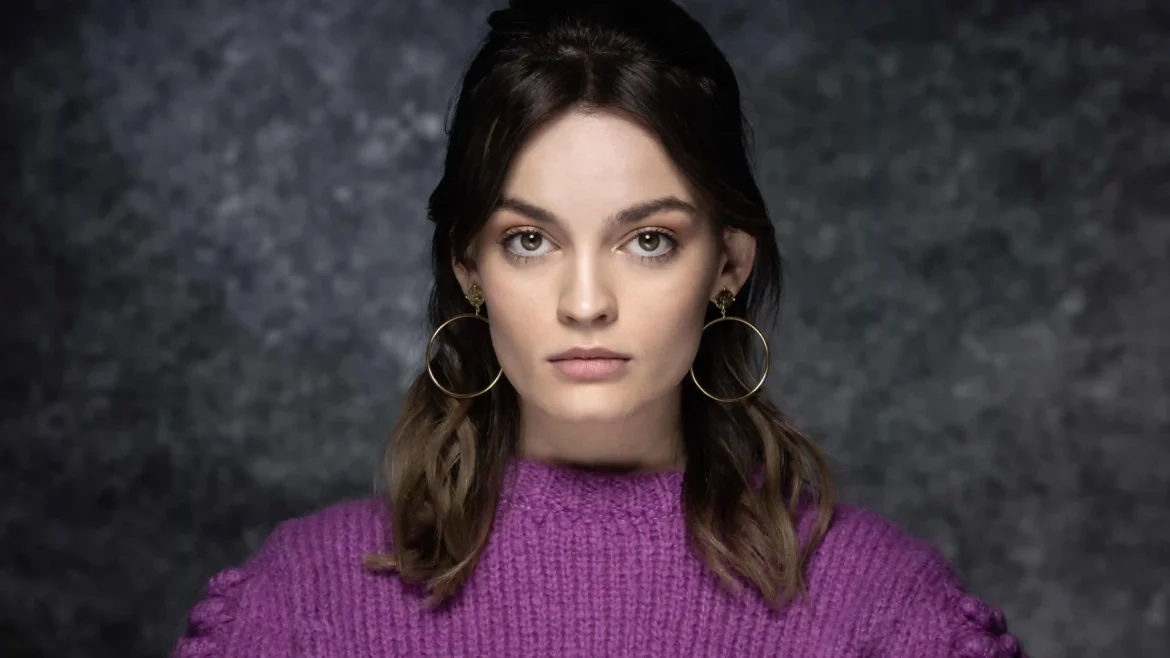 Studio portrait of actress Emma Mackey wearing a purple knit sweater and gold hoop earrings against a dark textured background in a 4K wallpaper
